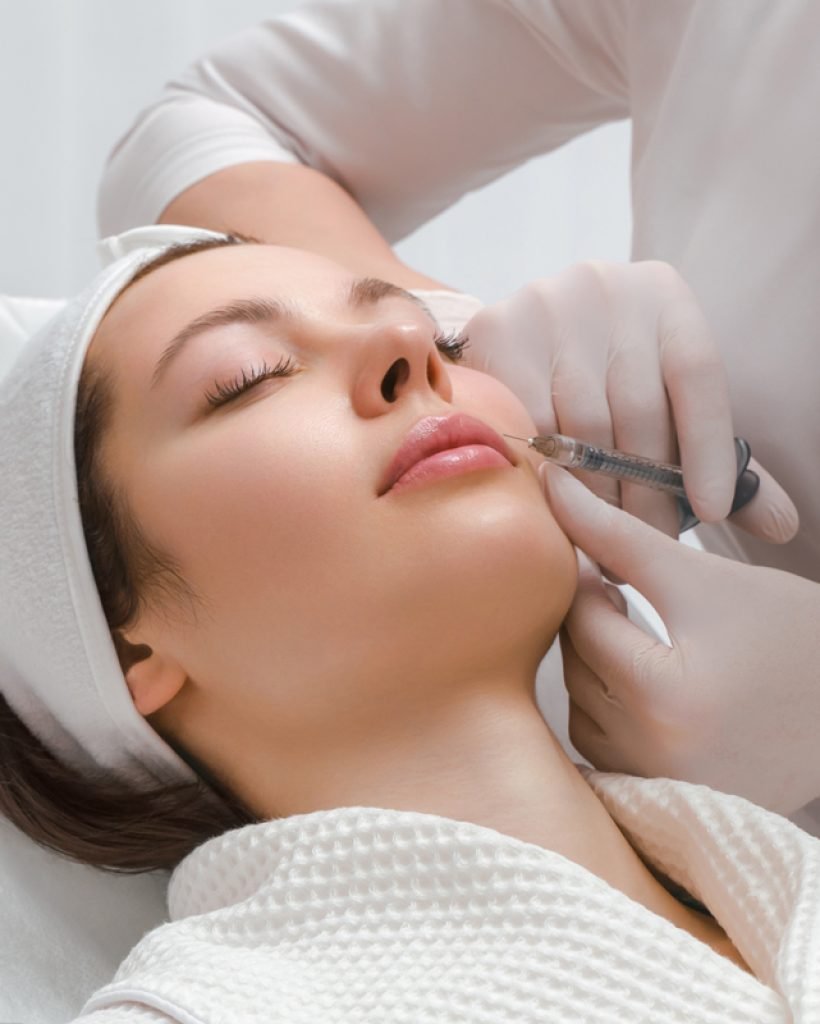 Paciente tumbada en camilla de consulta de medicina estética. Manos de doctor con jeringuilla preparada para administrar aumento de labios con ácido hialurónico